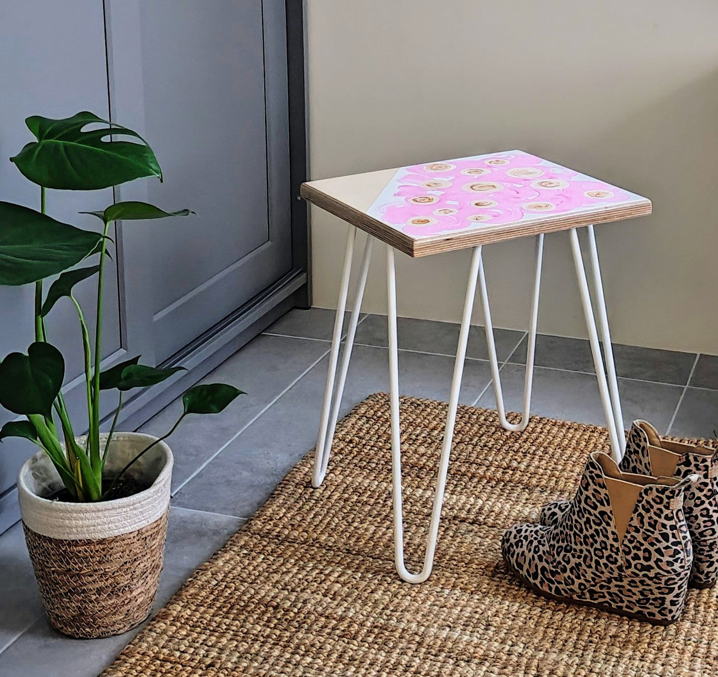 Plywood side table with hand-painted unique design in pink, white and gold. White metal hairpin legs. Side view. Next to the table is a pair of shoes and a plant.