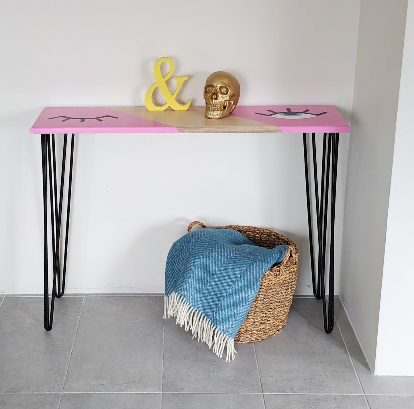 Front view of plywood console table painted with a pink background and a playful eye design. One eye is shut and the other one is open. The table has black hairpin legs.
Under the table is a basket with a blue blanket in it.