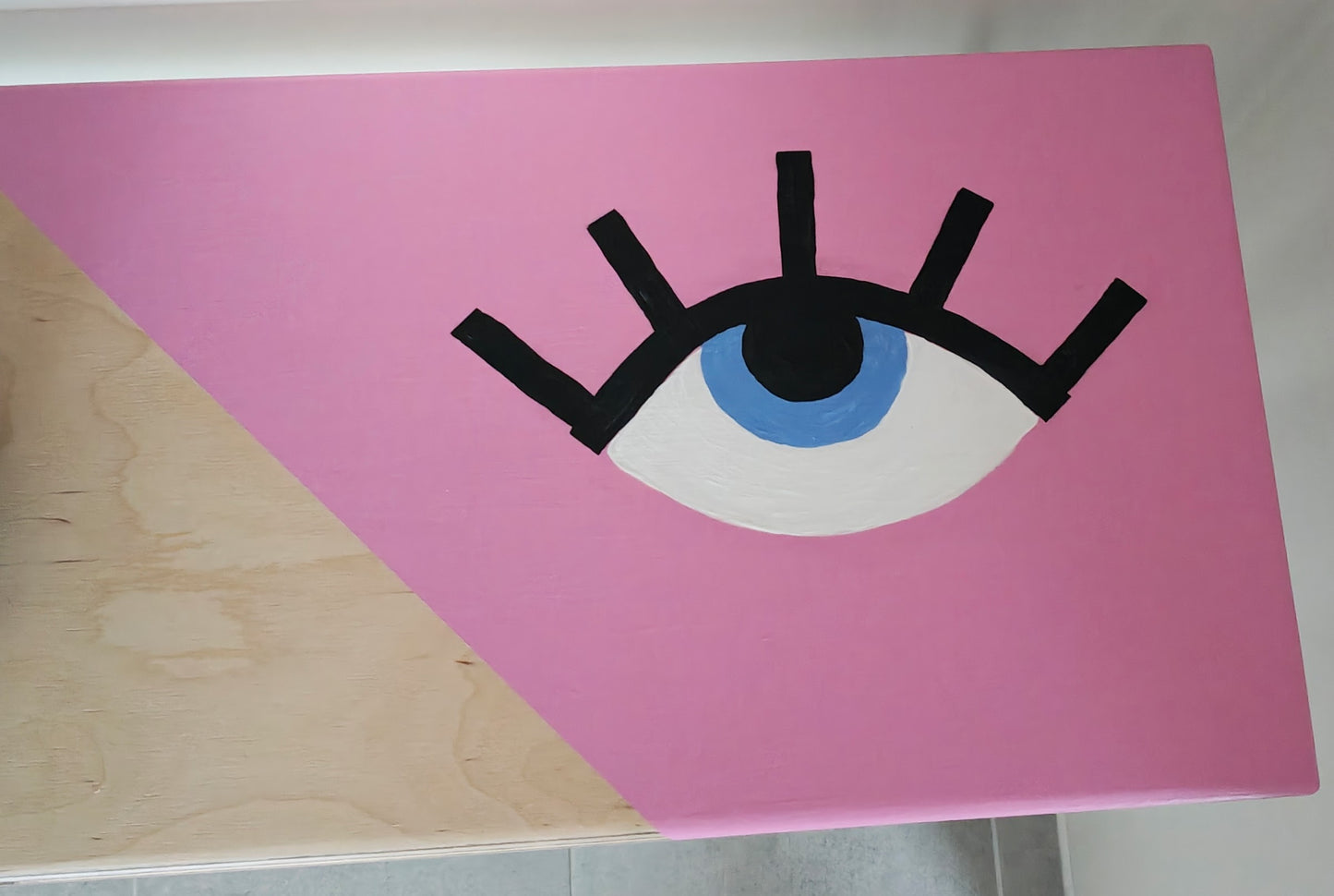 Close up view of plywood console table painted with a pink background and a playful eye design. The eye has a blue iris and long black eyelashes.