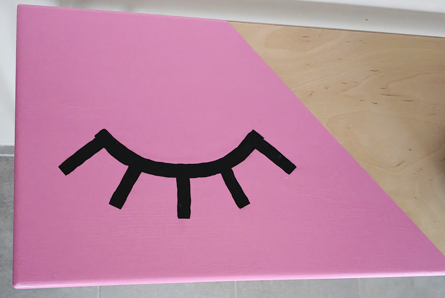 Close up view of plywood console table painted with a pink background and a playful eye design. The eye is closed and has long black eyelashes.