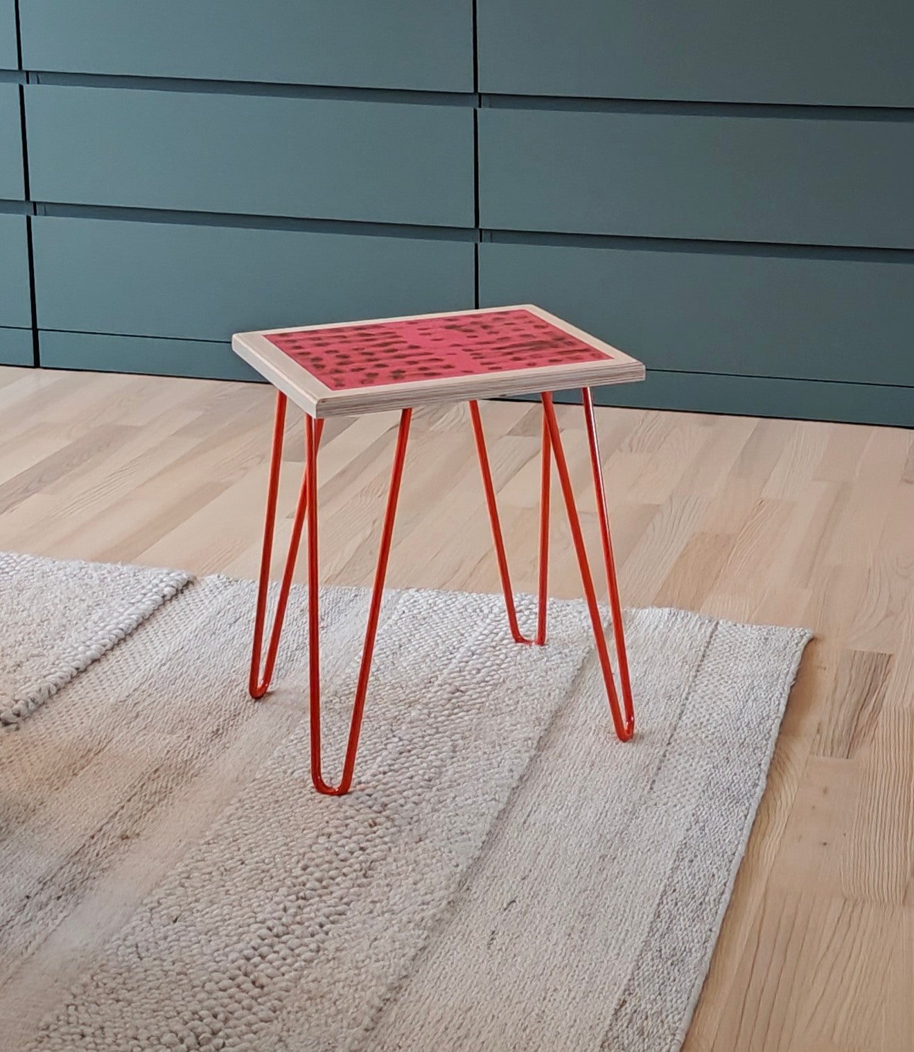 Plywood side table with hand-painted unique design in pink and gold. Orange metal hairpin legs. Side view. The table is standing on a white rug and there's a green chest of drawers in the background.