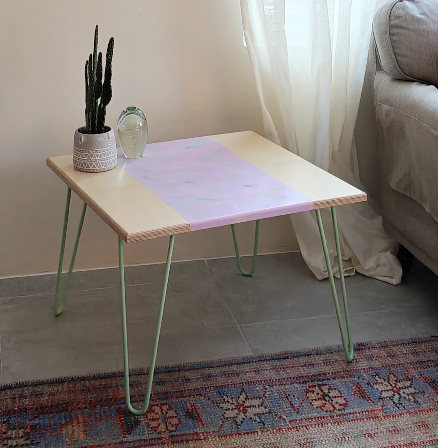 Plywood coffee table with handpainted unique design in purple and grey. Light green metal hairpin legs. On the table is a plant and a glass paperweight. The table is standing on a rug and there is a curtain and sofa in the background.