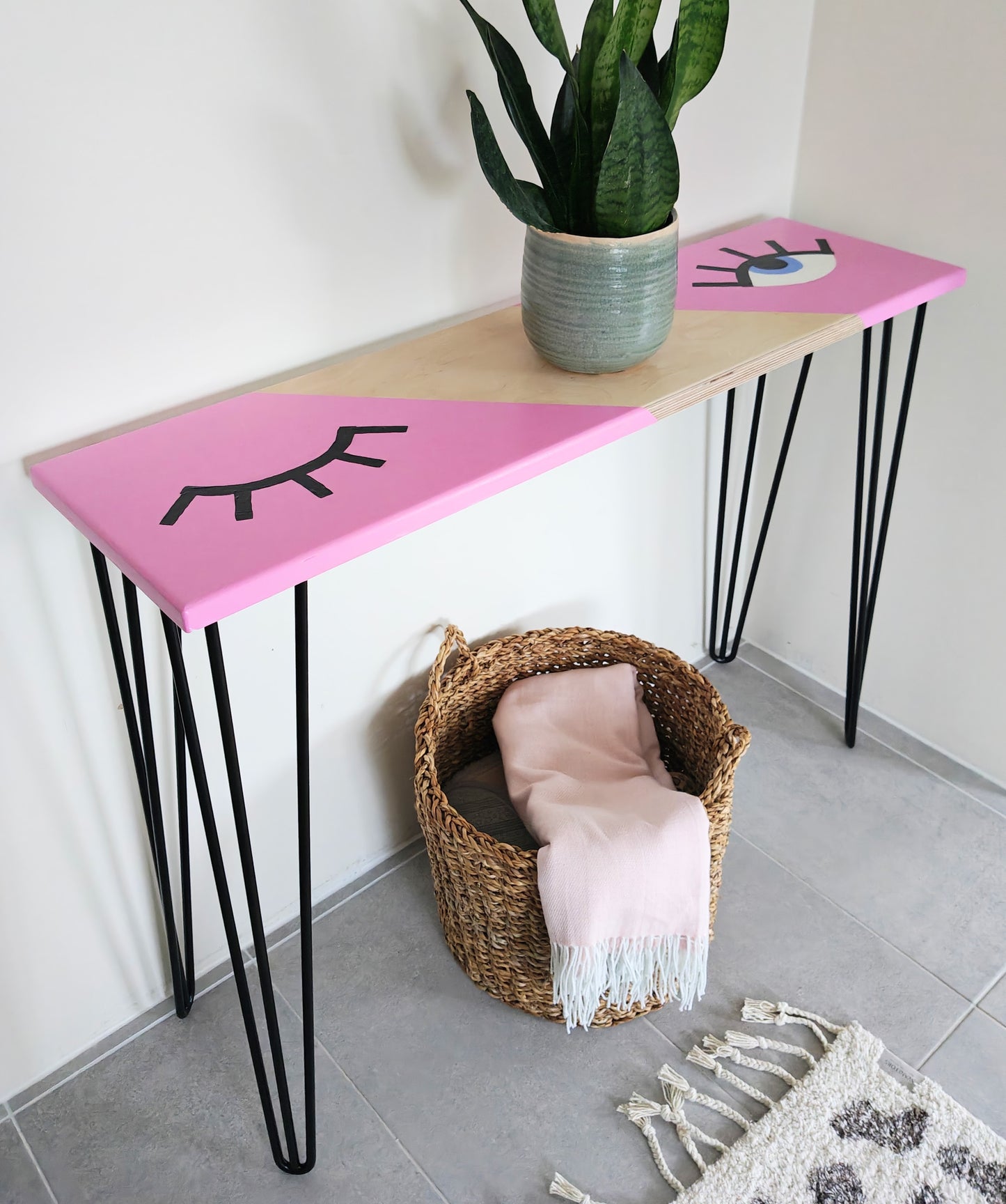 Side view of plywood console table painted with a pink background and a playful eye design. One eye is shut and the other one is open. The table has black hairpin legs.
Under the table is a basket with a pink blanket in it, and there is a plant pot on top of the table.