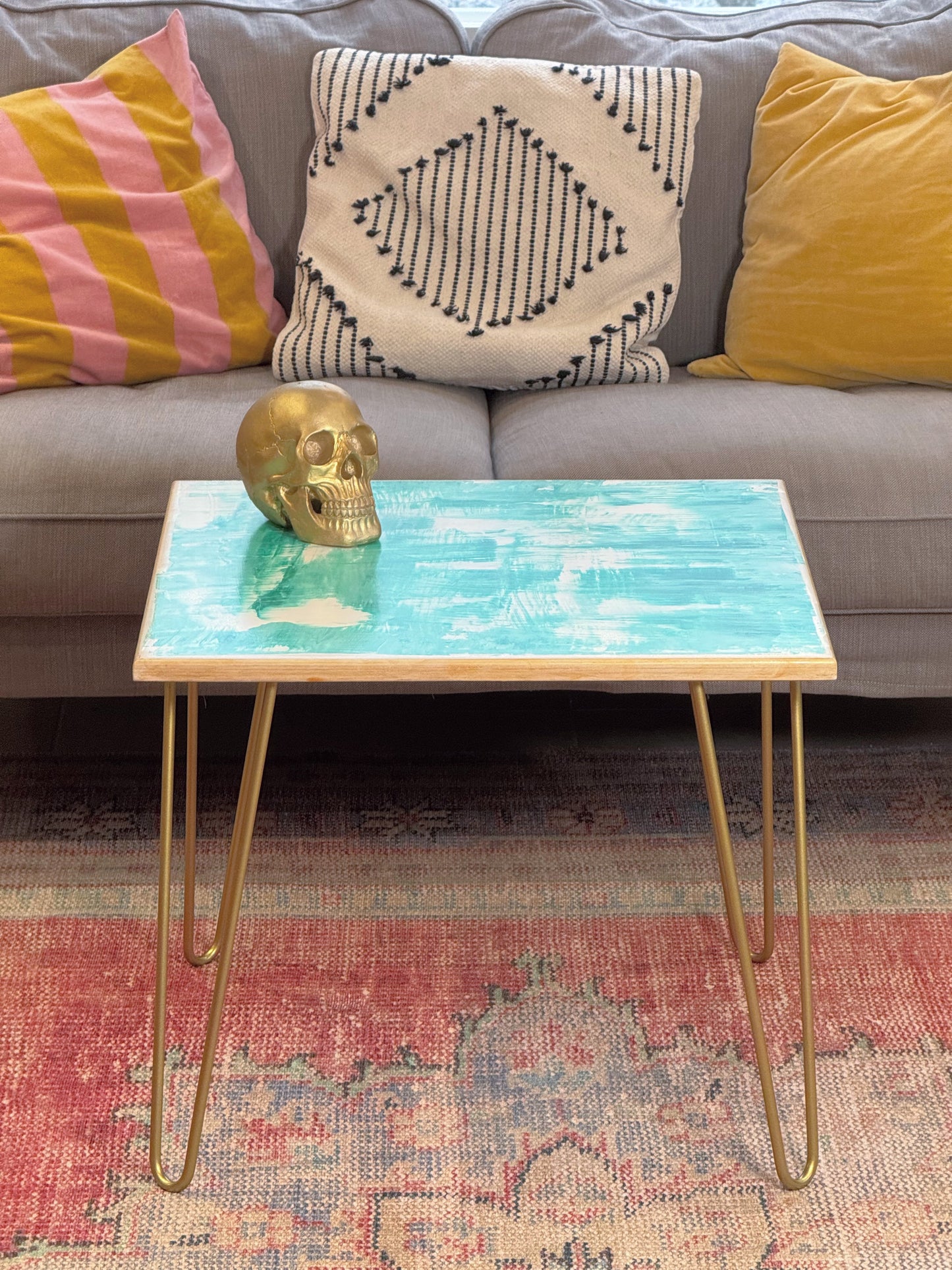 Plywood coffee table with hand-painted unique design in white, gold and green. Gold metal hairpin legs. Side view. There is a decorative golden skull on the table and a sofa with cushions in the background.