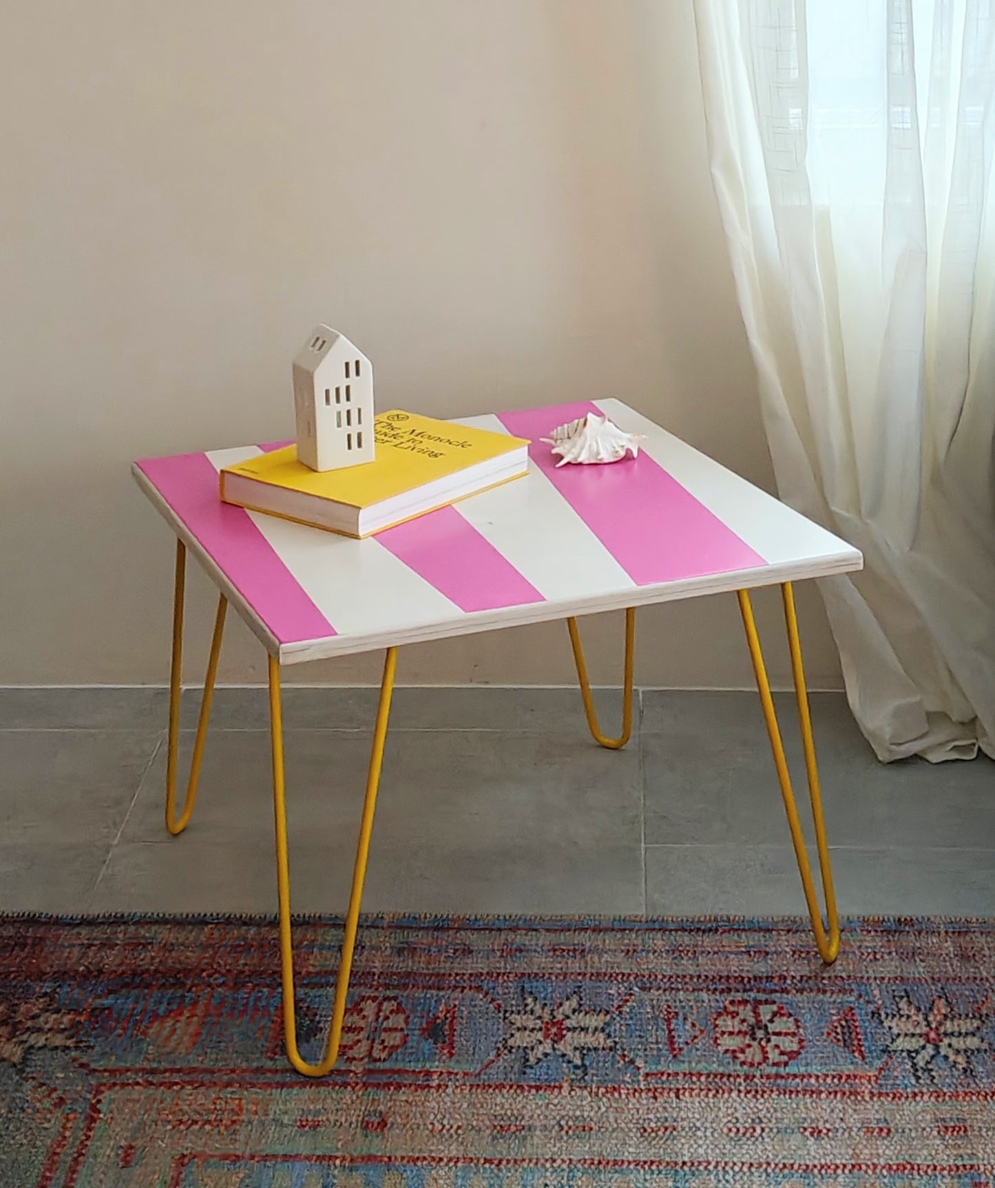 Plywood coffee table with handpainted unique design in pink and white. Bright yellow metal hairpin legs. Side view. On top of the table is a yellow book, small ceramic white house, and a white seashell. A curtain and a rug are visible in the background.