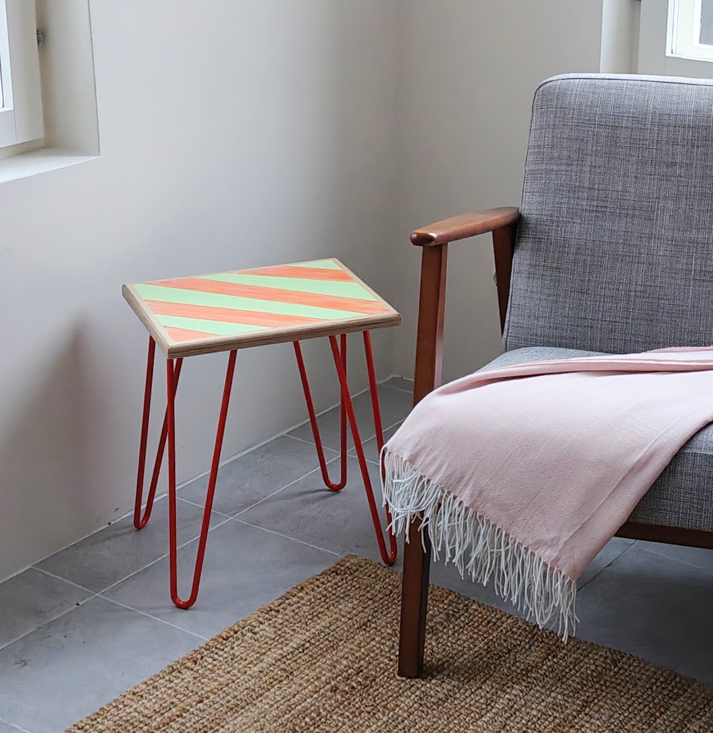 Plywood side table with hand-painted unique design in orange and green. Orange metal hairpin legs. Side view. The table is next to a grey armchair with a pink throw.