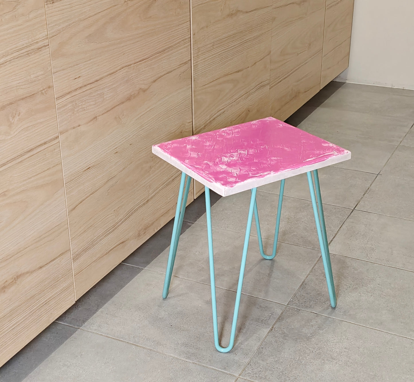 Plywood side table with hand-painted unique design in pink and white. Light blue metal hairpin legs. Side view
