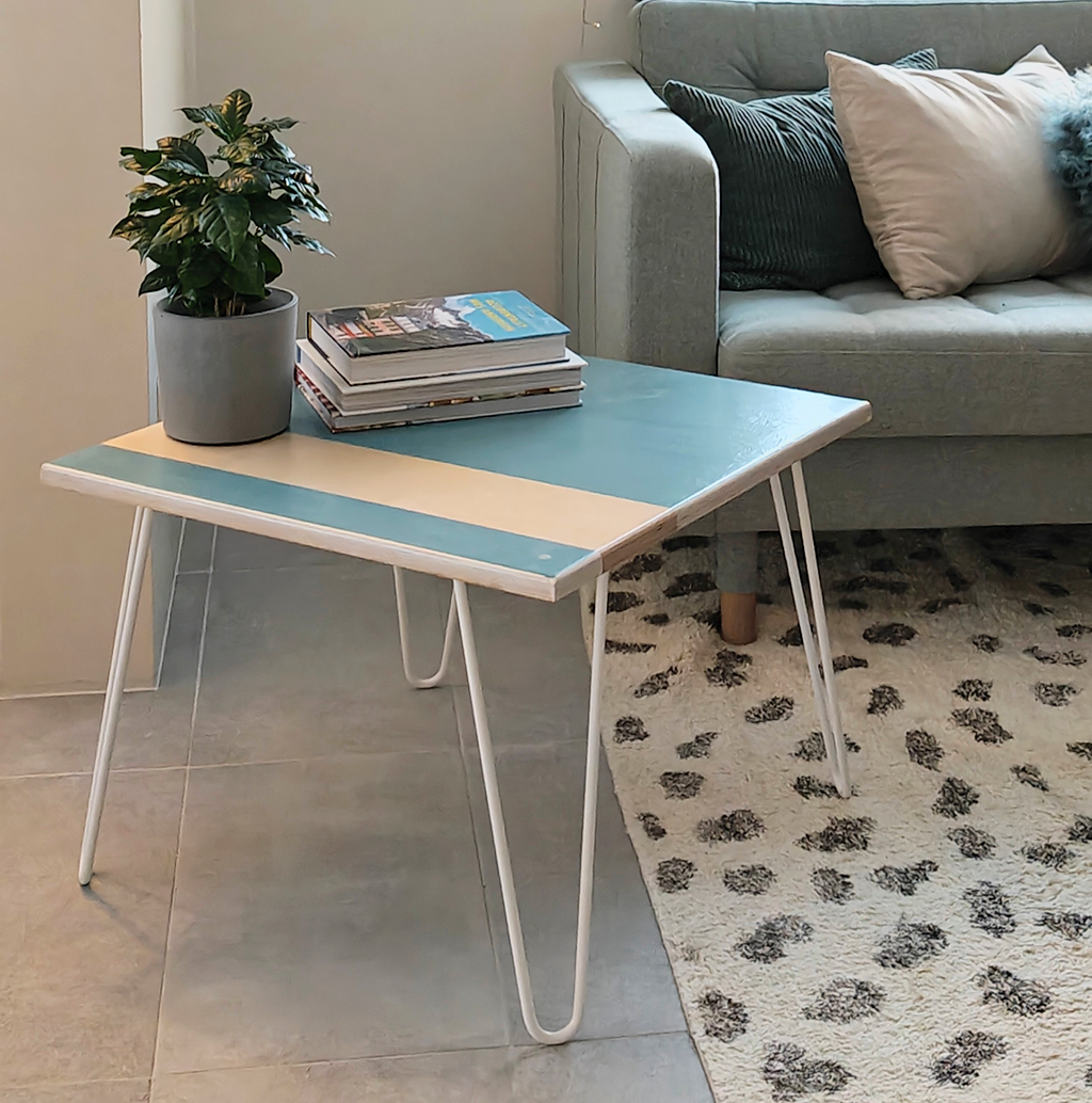 Plywood coffee table with handpainted unique design in blue and white. White metal hairpin legs. Side view. On the table is a plant and a pile of books. Behind the table is a light green sofa with cushions on. The table is standing on a spotted black and white rug.