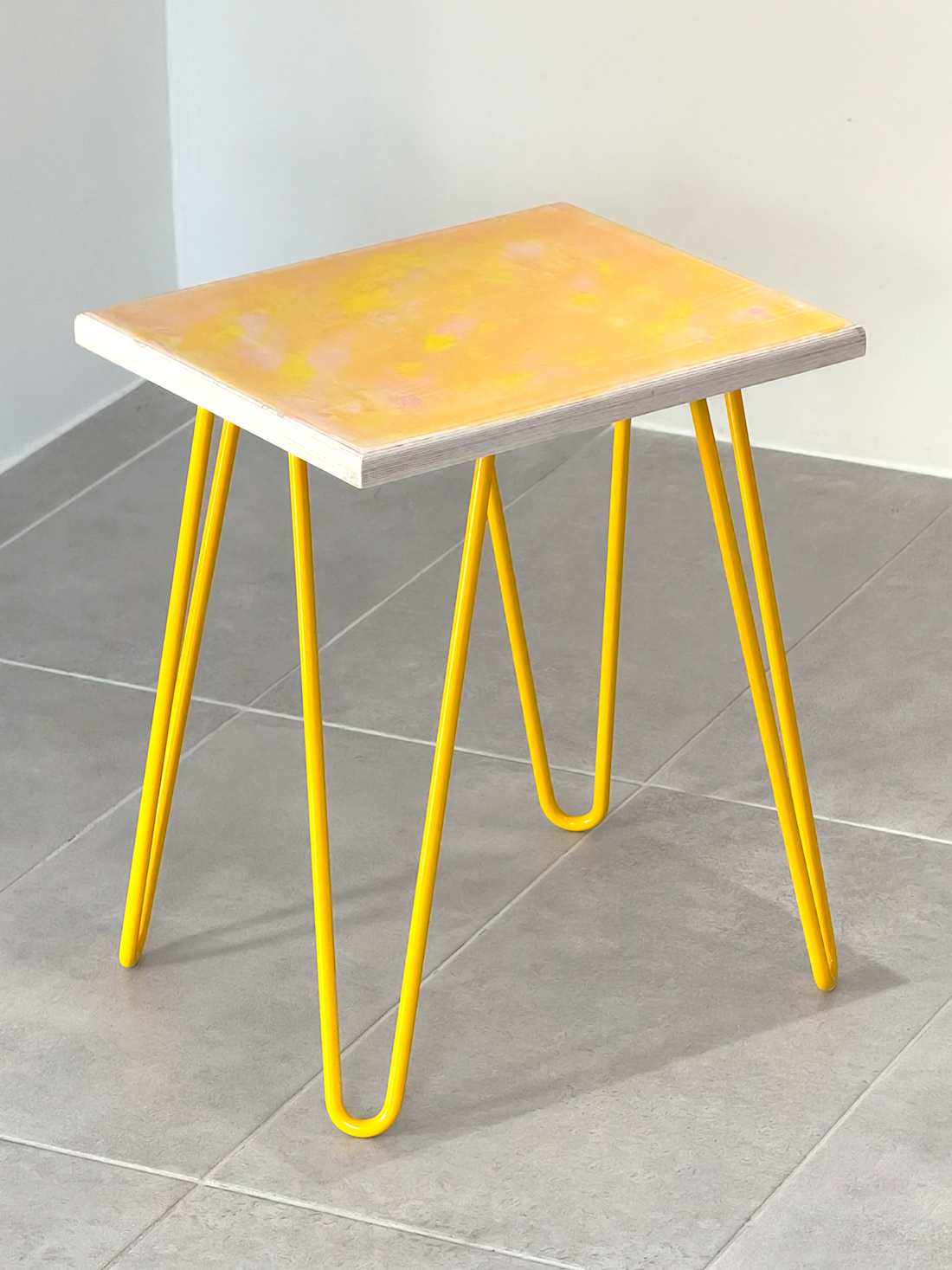 Plywood side table with hand-painted unique design in pink and yellow. Yellow metal hairpin legs. Side view