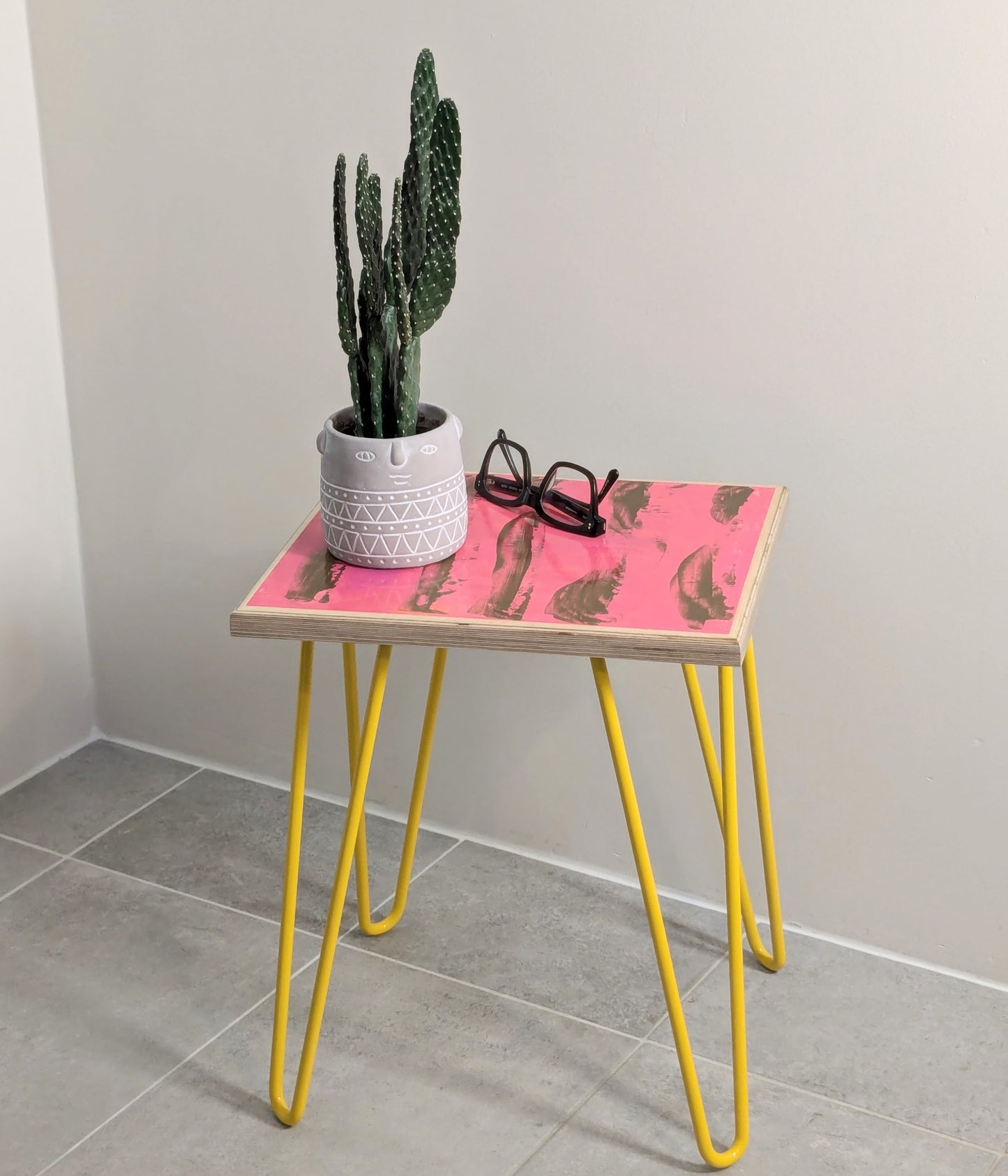 Plywood side table with hand-painted unique design in pink and gold. Yellow metal hairpin legs. Side view. Potted cactus and a pair of glasses are on the table.