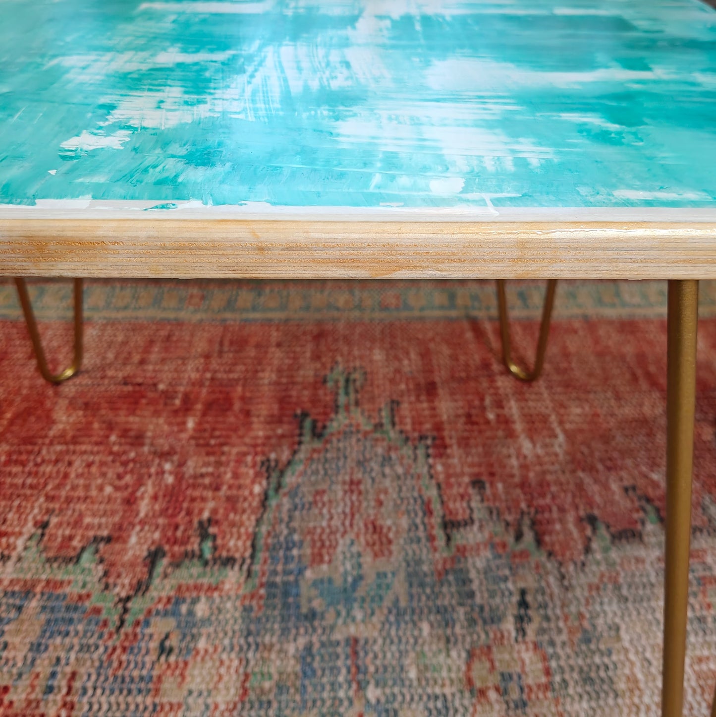 Plywood coffee table with hand-painted unique design in white, gold and green. Gold metal hairpin legs. Close up side view.