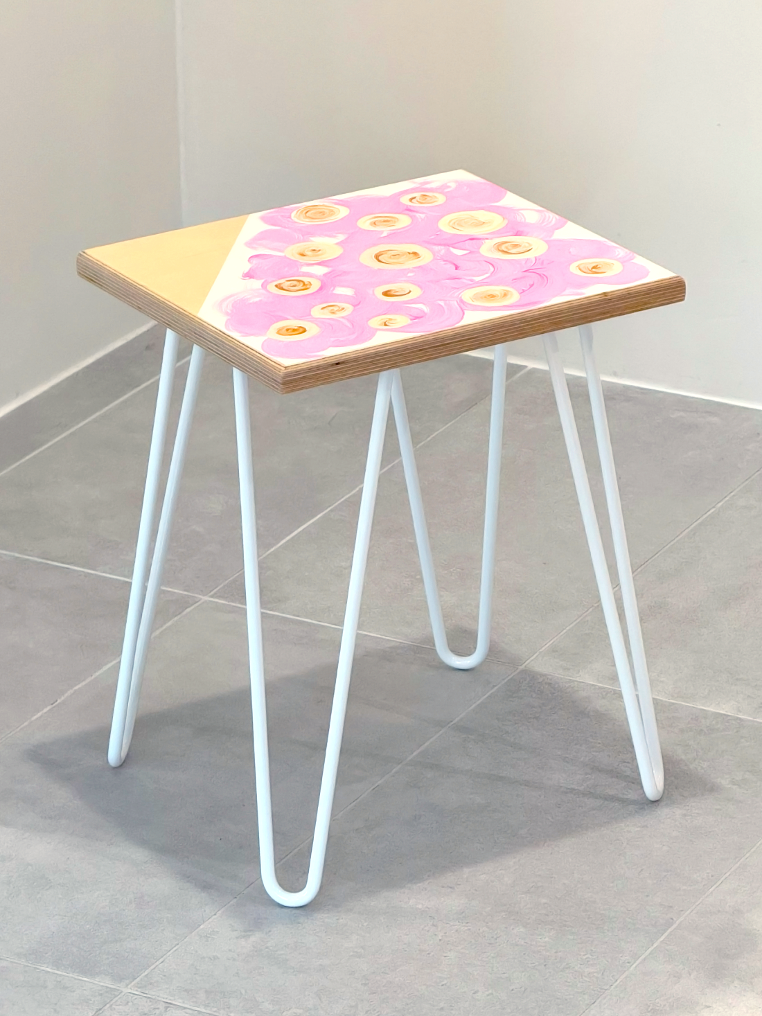 Plywood side table with hand-painted unique design in pink, white and gold. White metal hairpin legs. Side view.
