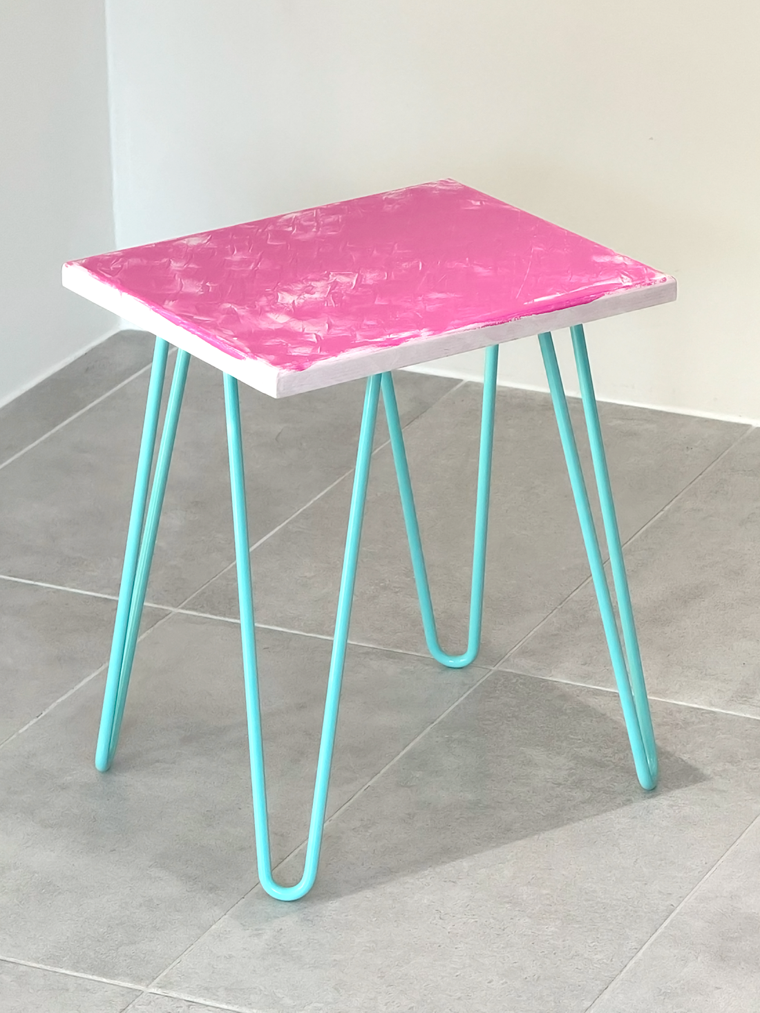 Plywood side table with hand-painted unique design in pink and white. Light blue metal hairpin legs. Side view